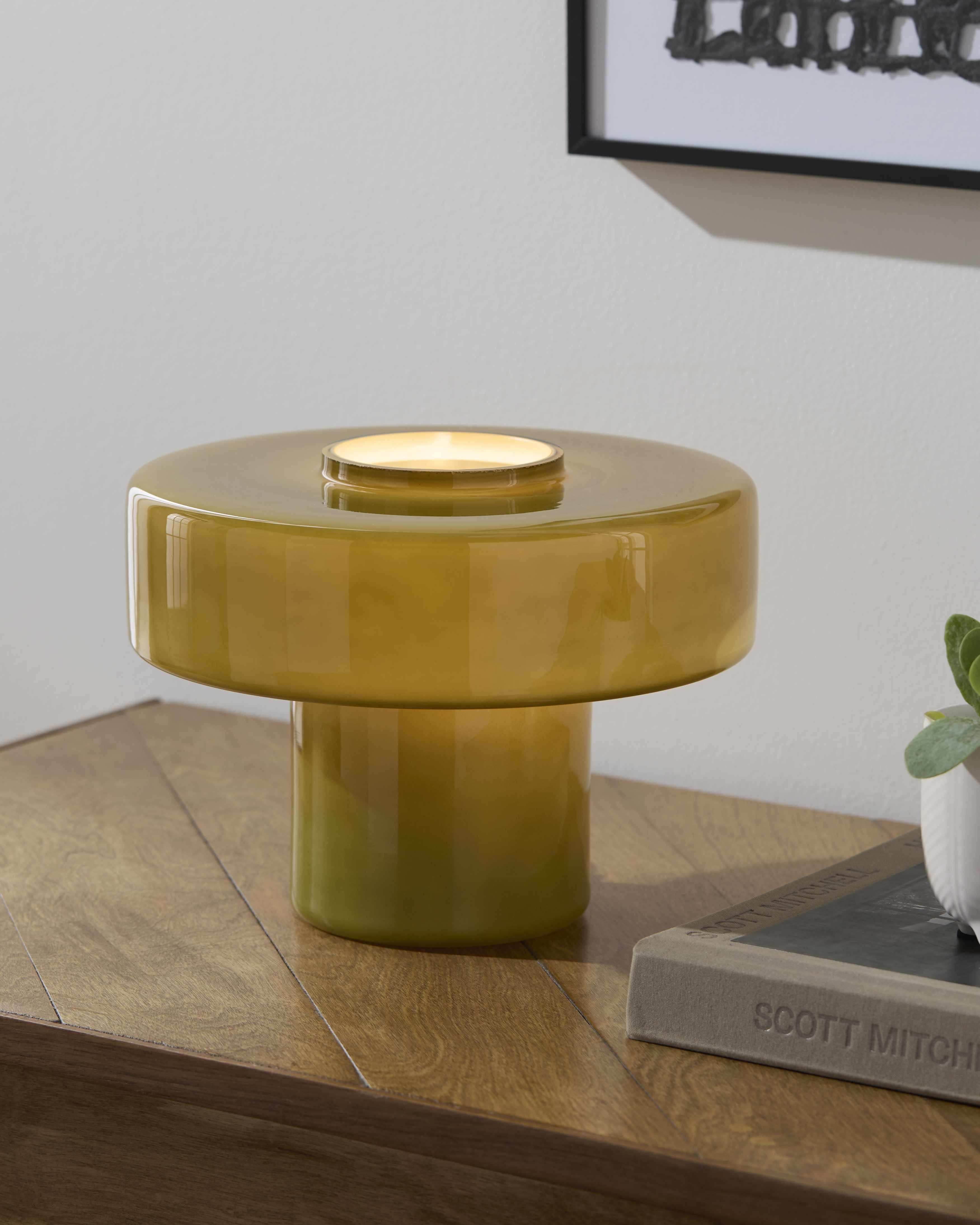 Round yellow contemporary table lamp on a wooden surface with a book and plant in the background.